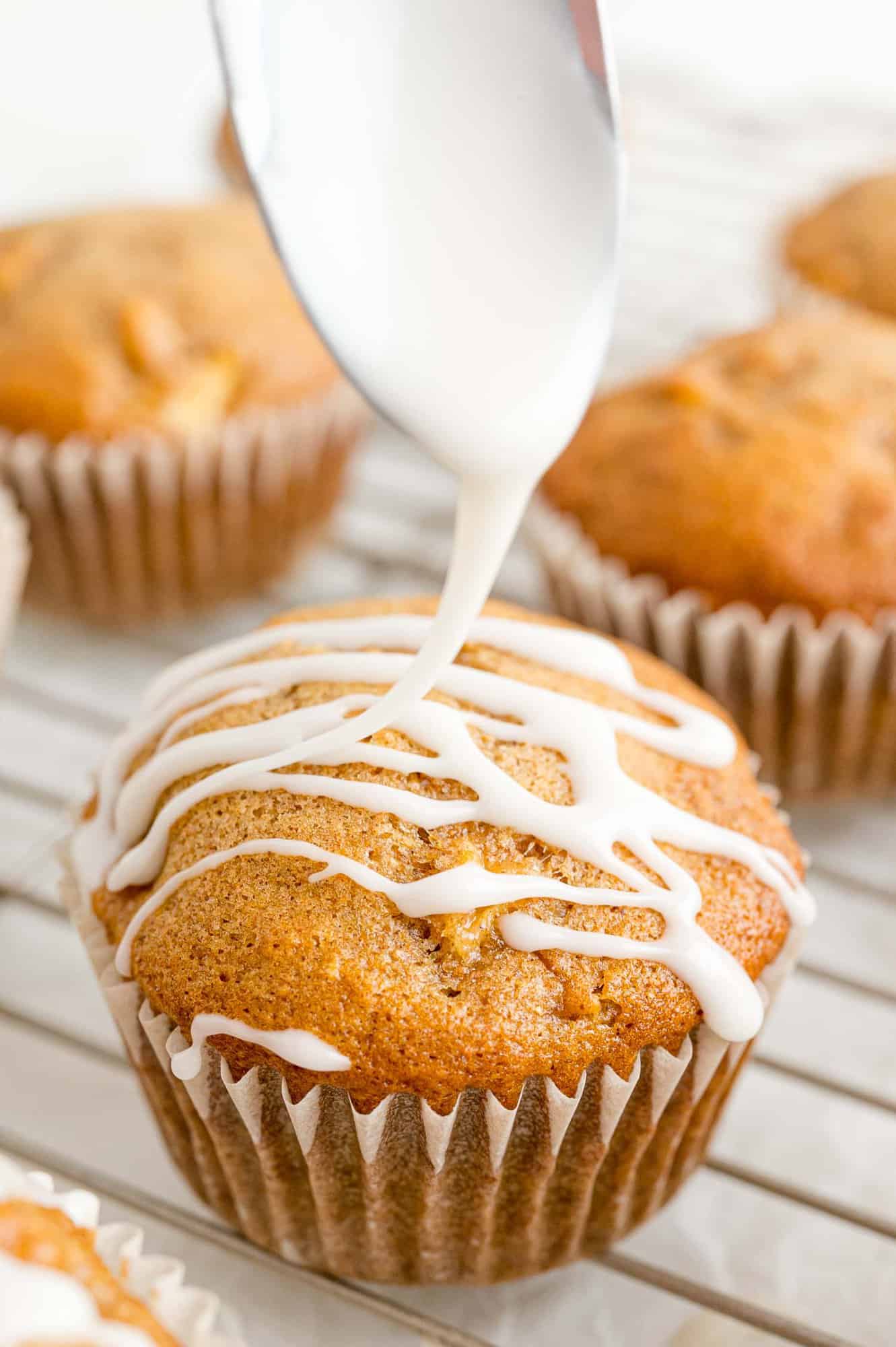 A spoon drizzling glaze over an apple cider muffin on a wire rack.