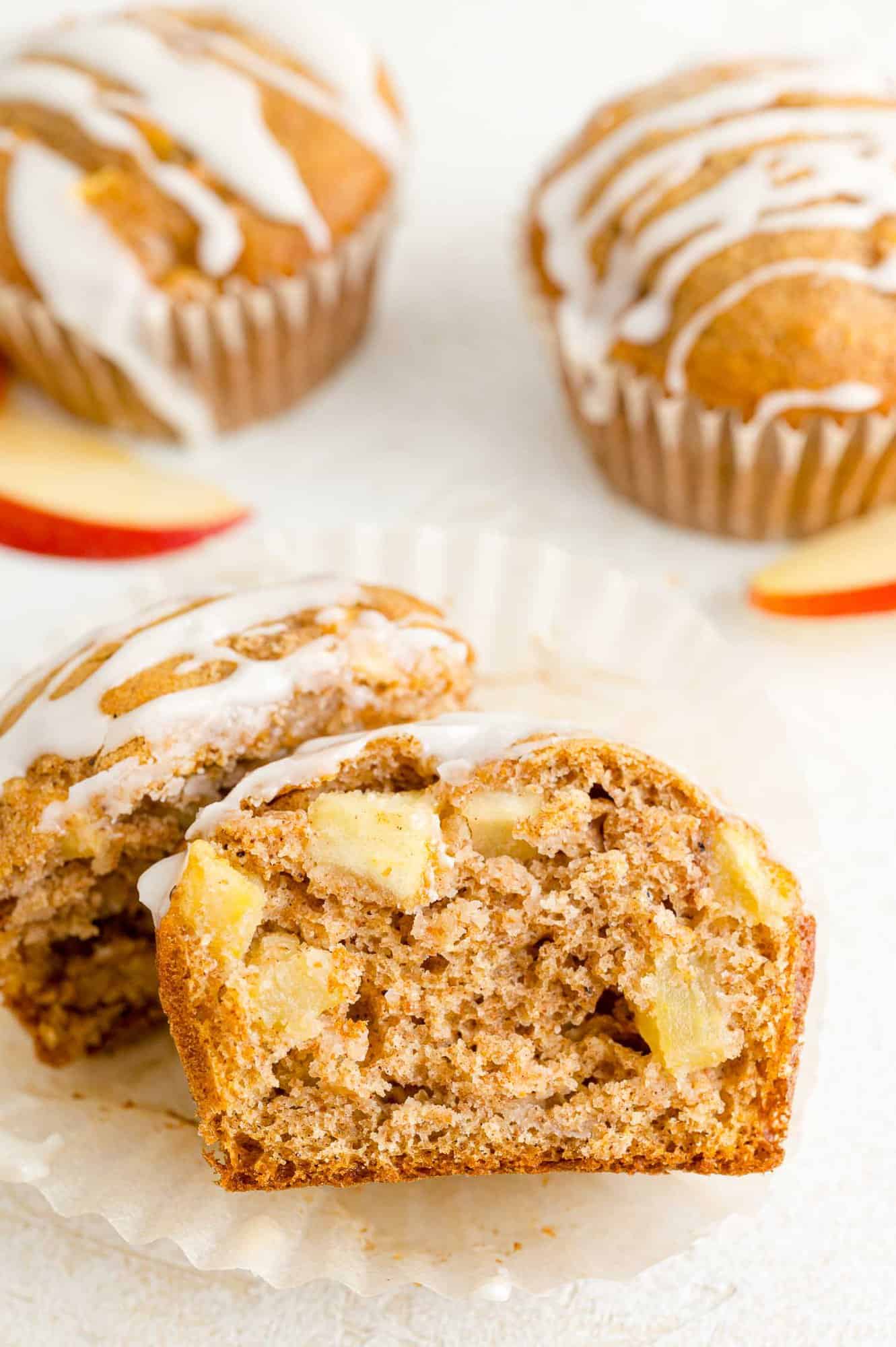 A glazed apple cider muffin cut in half to reveal chunks of apples inside, with more muffins in the background.