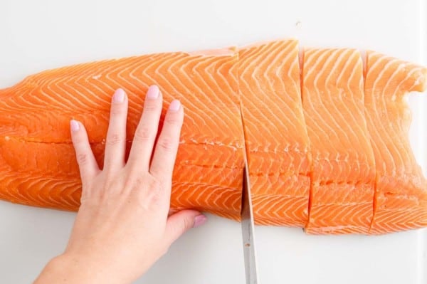 A hand holds a side of salmon in place while the other hand uses a sharp knife to cut the salmon into filets.