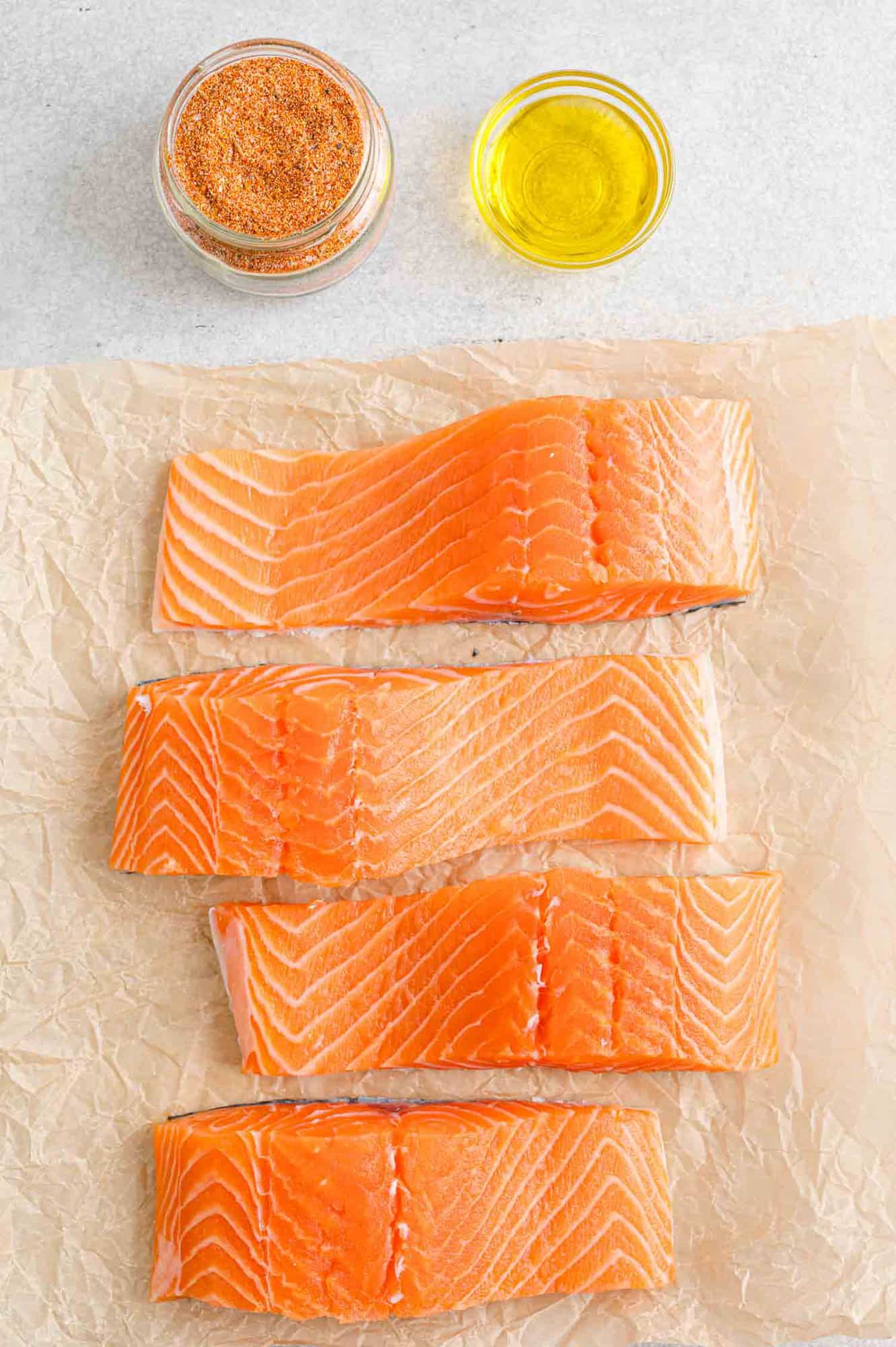 Four salmon filets on a sheet of parchment paper next to small bowls of olive oil and salmon seasoning.