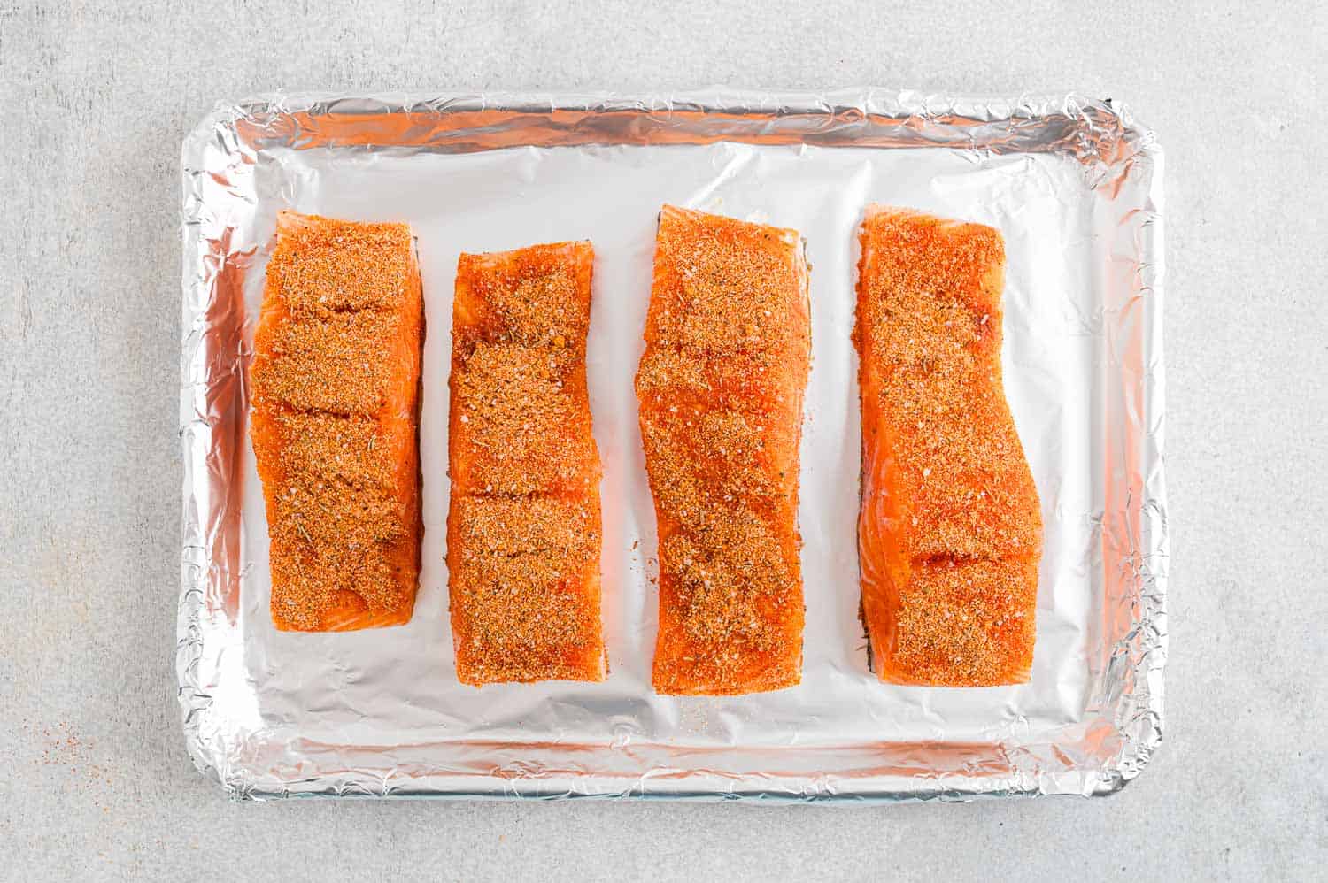 Four seasoned salmon filets on a foil-lined baking sheet.