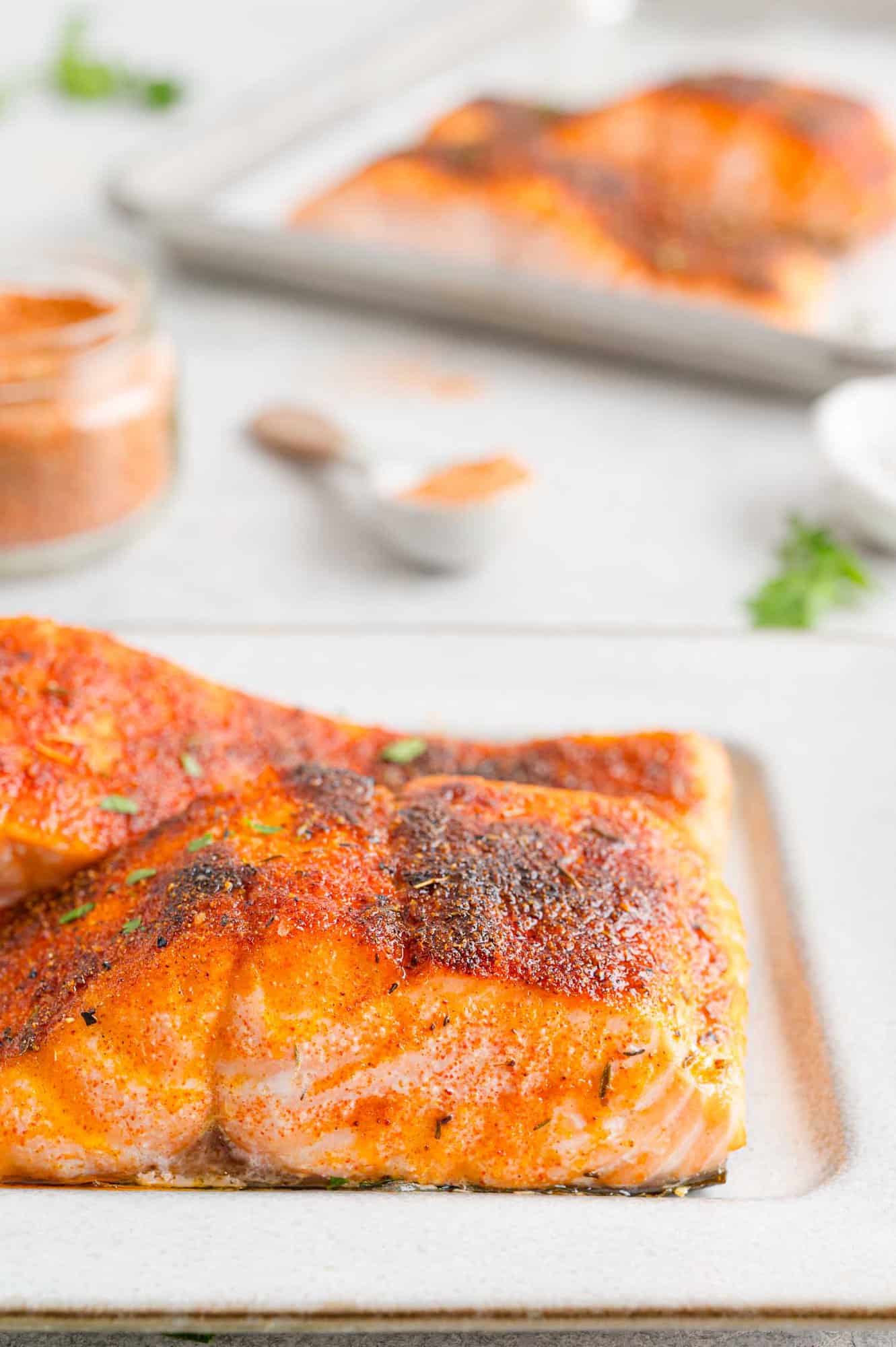 Close up side view of broiled salmon filets on a plate, with more filets on a foil-lined baking sheet in the background.