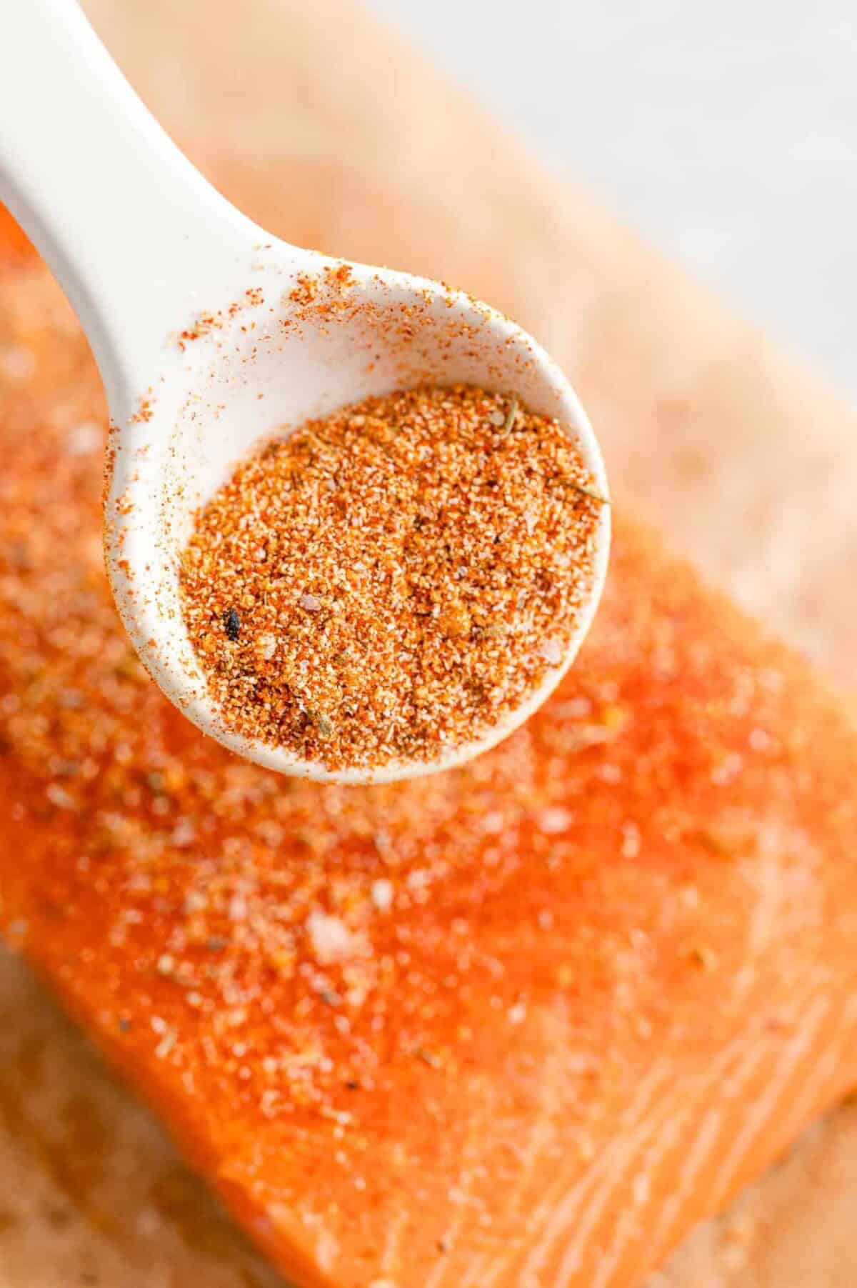 A white spoon holds a blend of dry seasoning above a raw salmon fillet, which is sprinkled with the same seasoning mix.