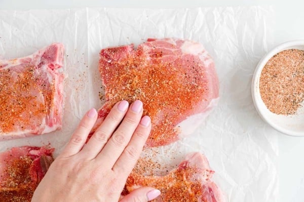 A hand pats seasoning rub onto uncooked pork chops.