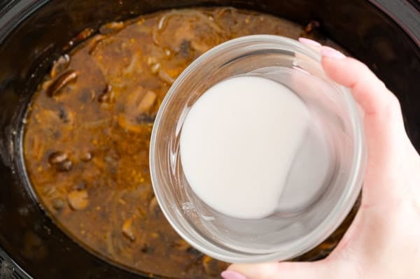 A bowl with a cornstarch slurry is held over mushroom and onion gravy inside the slow cooker.