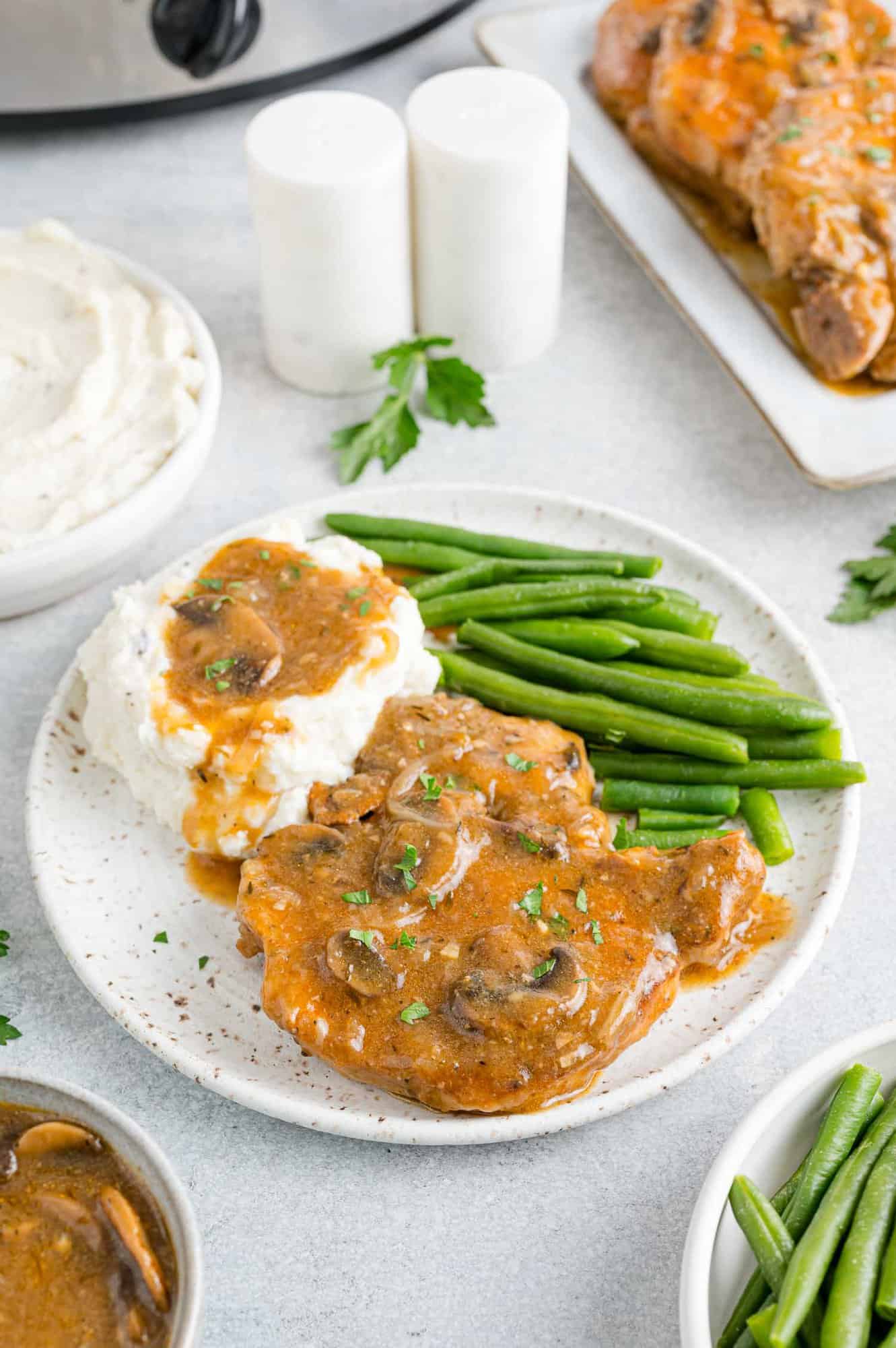 Slow cooker pork chops served on a plate with gravy, mashed potatoes, and green beans, next to dishes of potatoes and beans on a countertop.