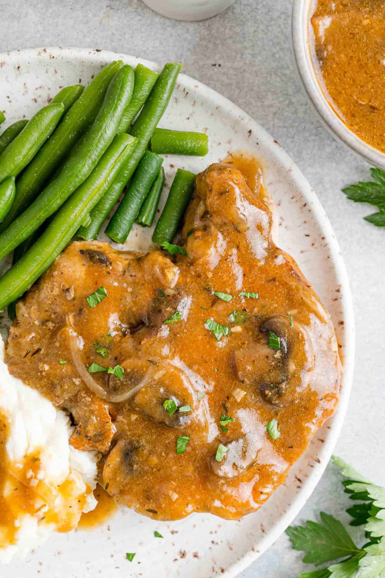 Overhead view of slow cooker pork chops served on a plate, covered in gravy next to mashed potatoes and green beans.