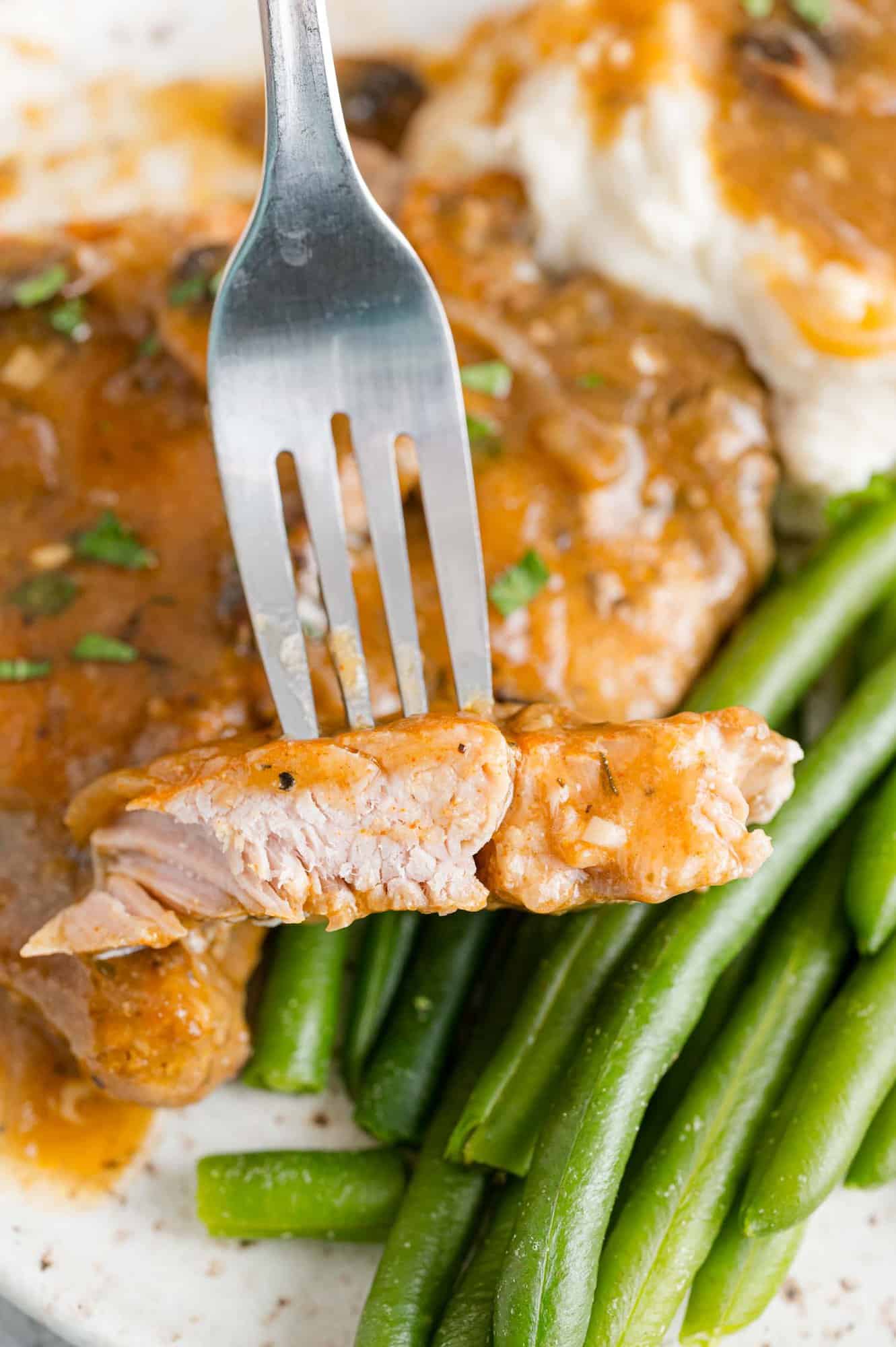 Close up of a piece of pork chop on a fork, held above a plate of pork chops, mashed potatoes, and green beans.