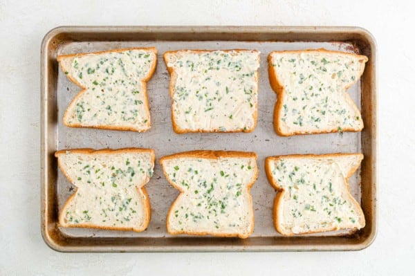 Six slices of Texas toast spread with garlic butter arranged on a baking sheet.
