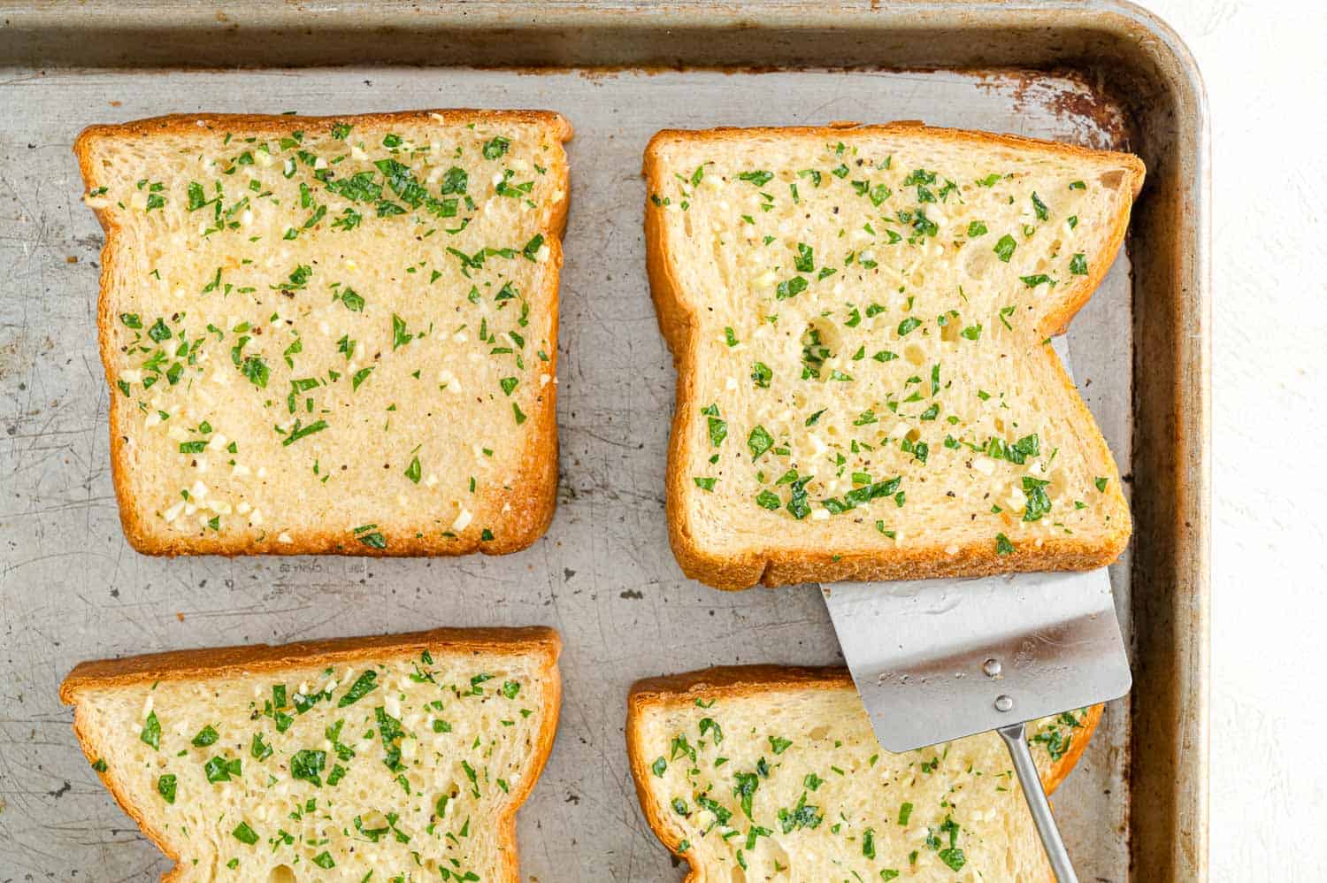 A spatula is used to flip slices of Texas toast garlic bread on a baking sheet.