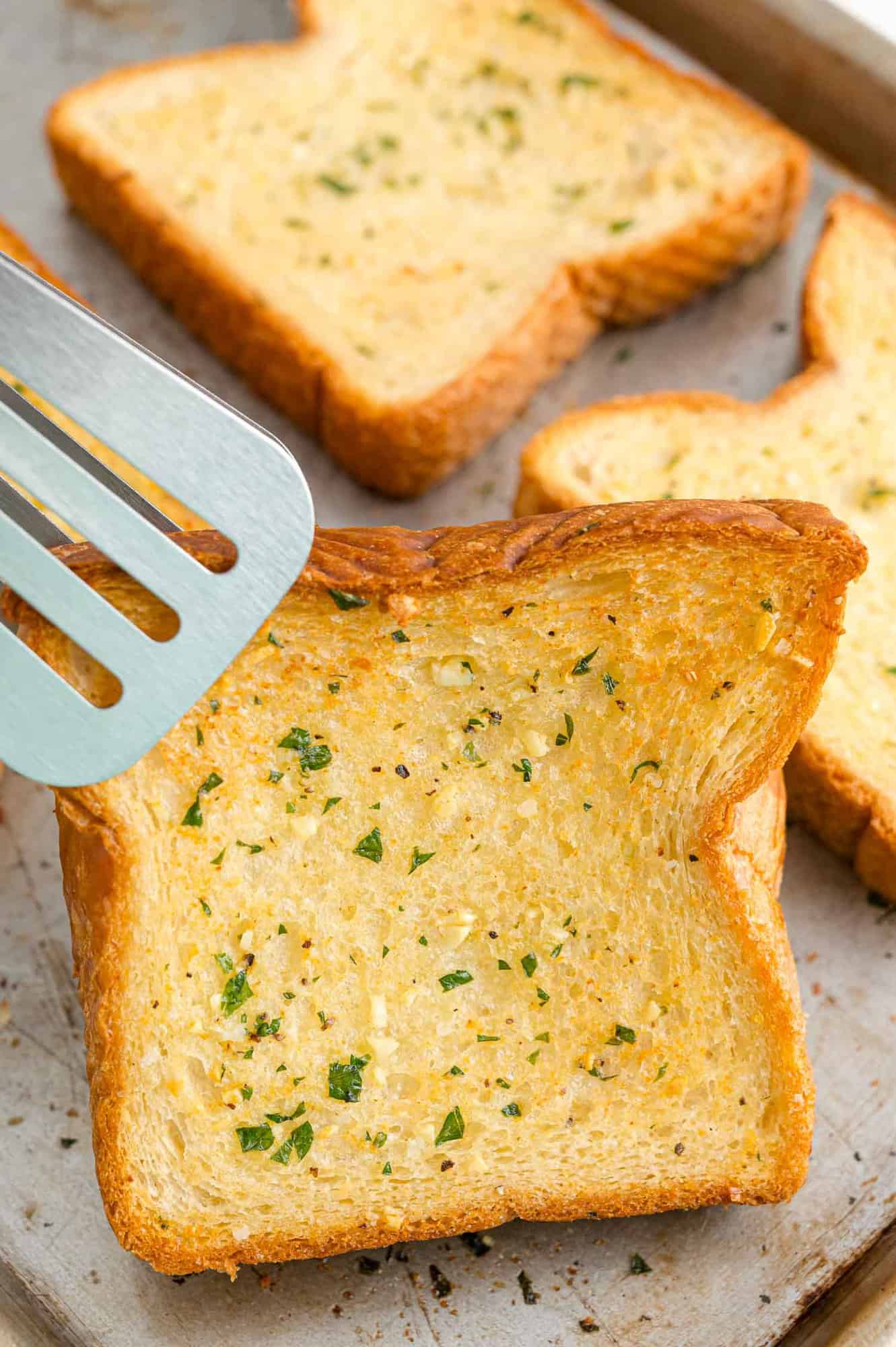 A pair of tongs picks up a slice of Texas toast garlic bread from a baking sheet.