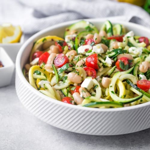Zucchini noodle salad in white bowl with tomatoes, feta, and beans.