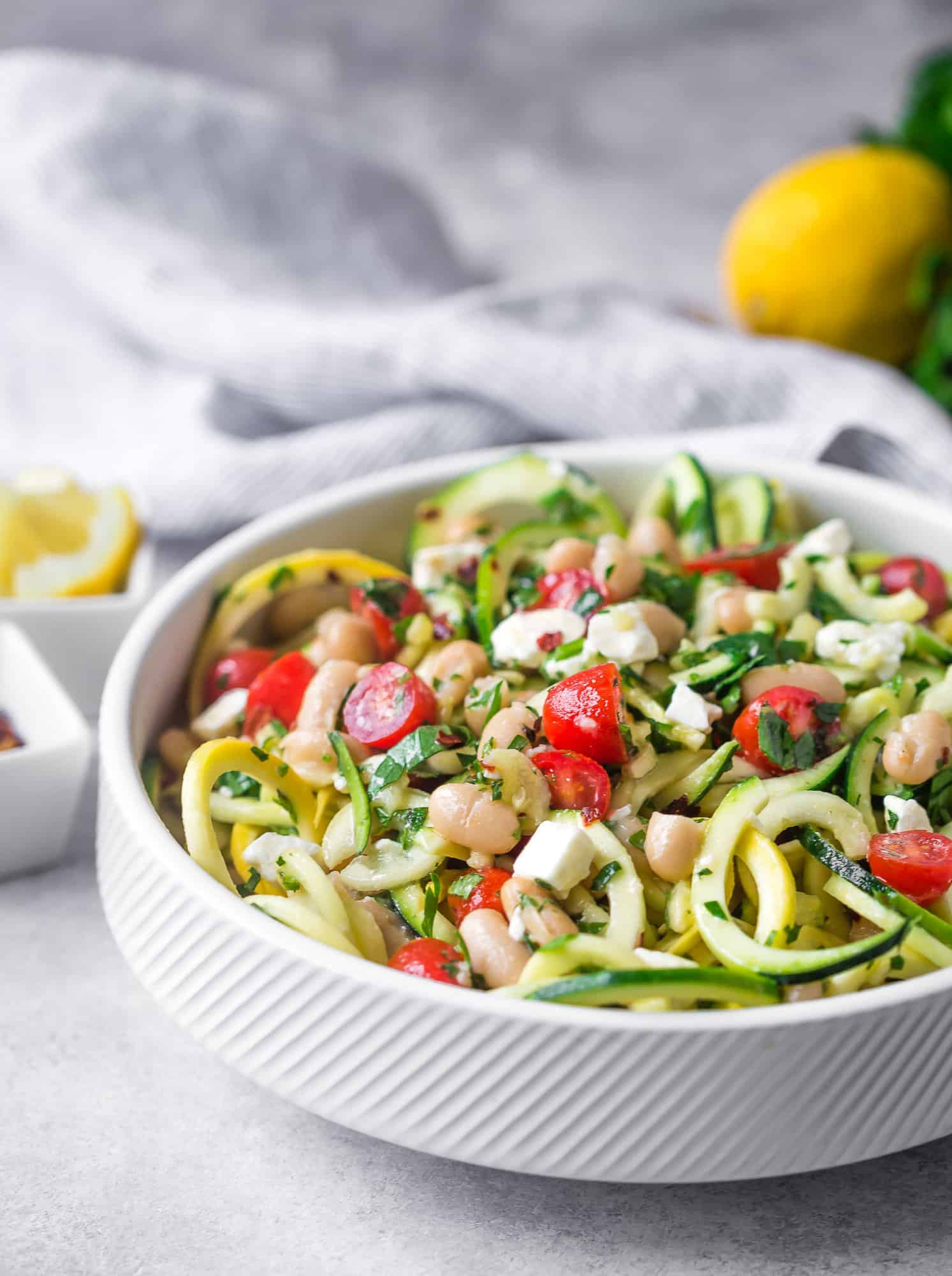 Zucchini noodle salad in a white bowl with tomatoes, feta, beans.