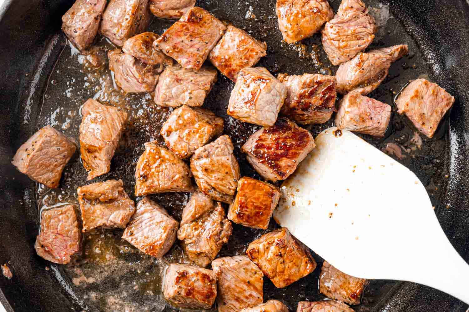 Fully cooked steak bites in a pan, being stirred with white spatula.