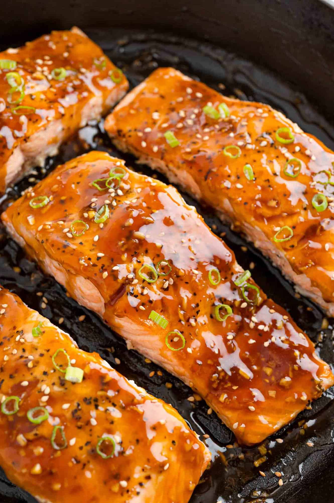 Fillets of teriyaki salmon in a black frying pan, garnished with sesame and green onion.