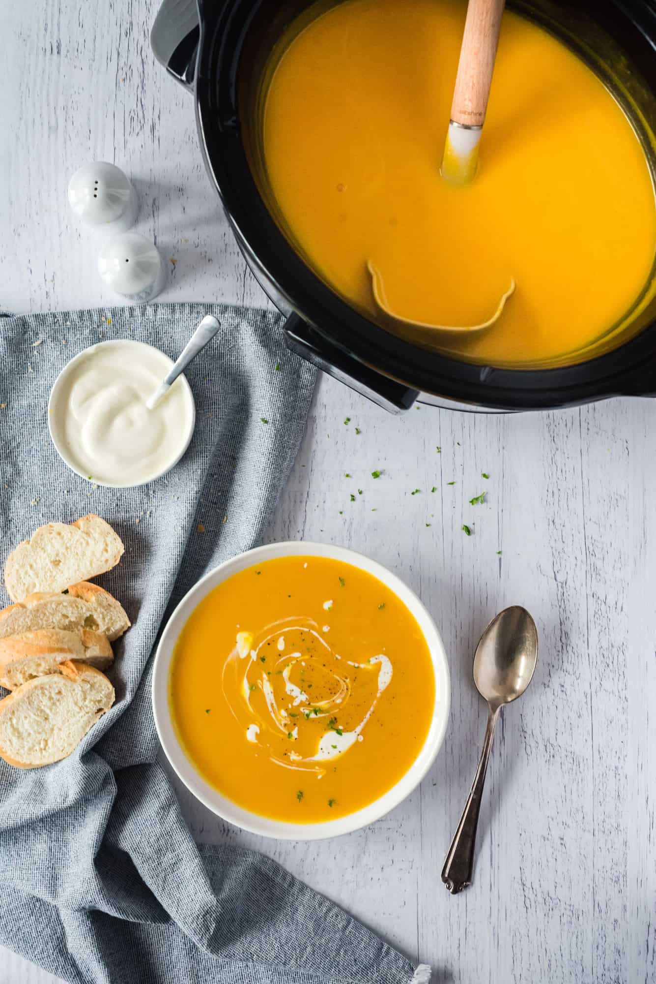 Overhead view of a bowl of pumpkin soup on a countertop next to a crockpot and slices of bread.