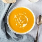 Overhead view of a bowl of pumpkin soup swirled with cream, next to bread slices and a spoon.