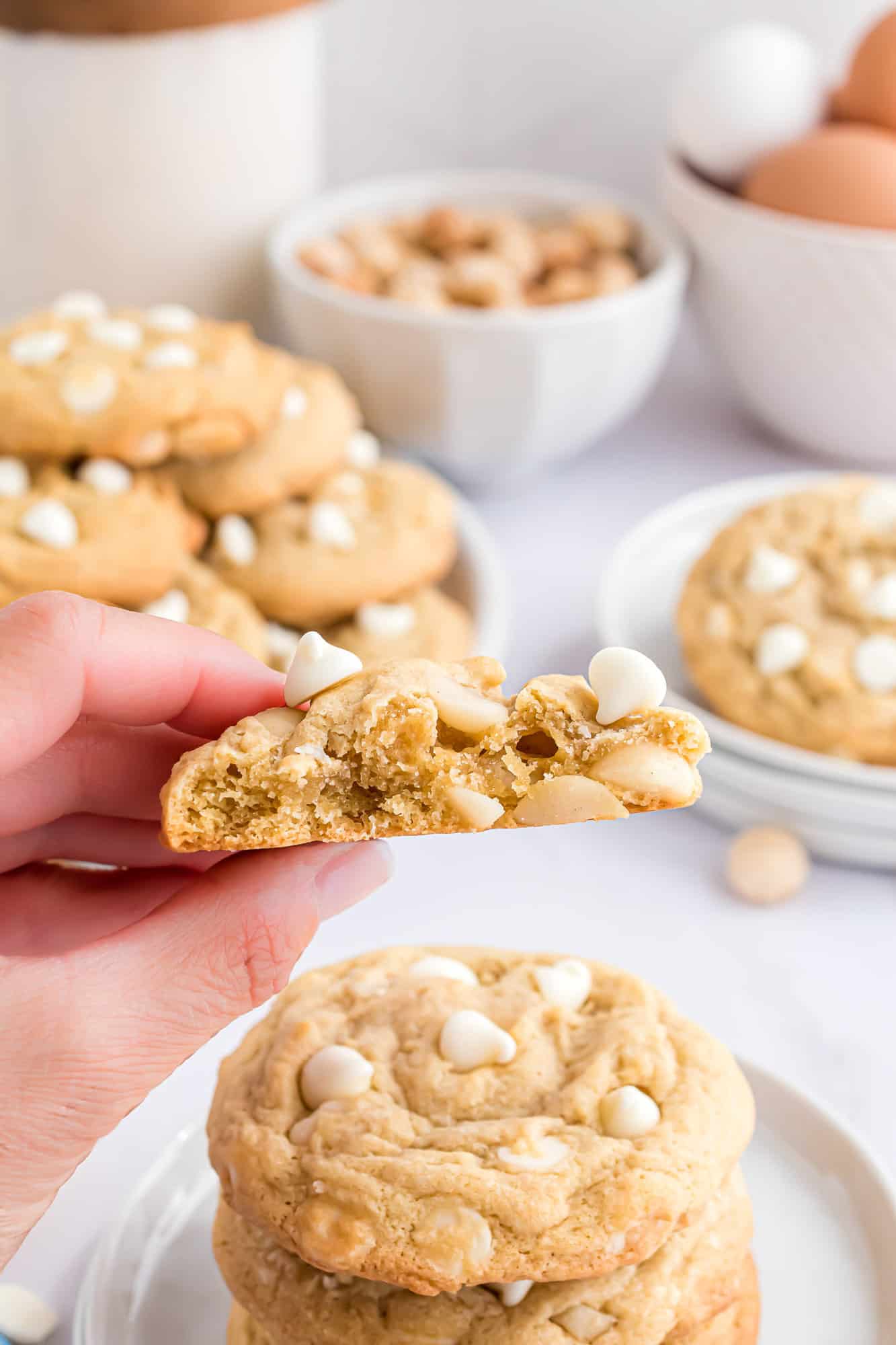 A hand holding up one half of a white chocolate macadamia nut cookie, with more cookies on a plate in the background.