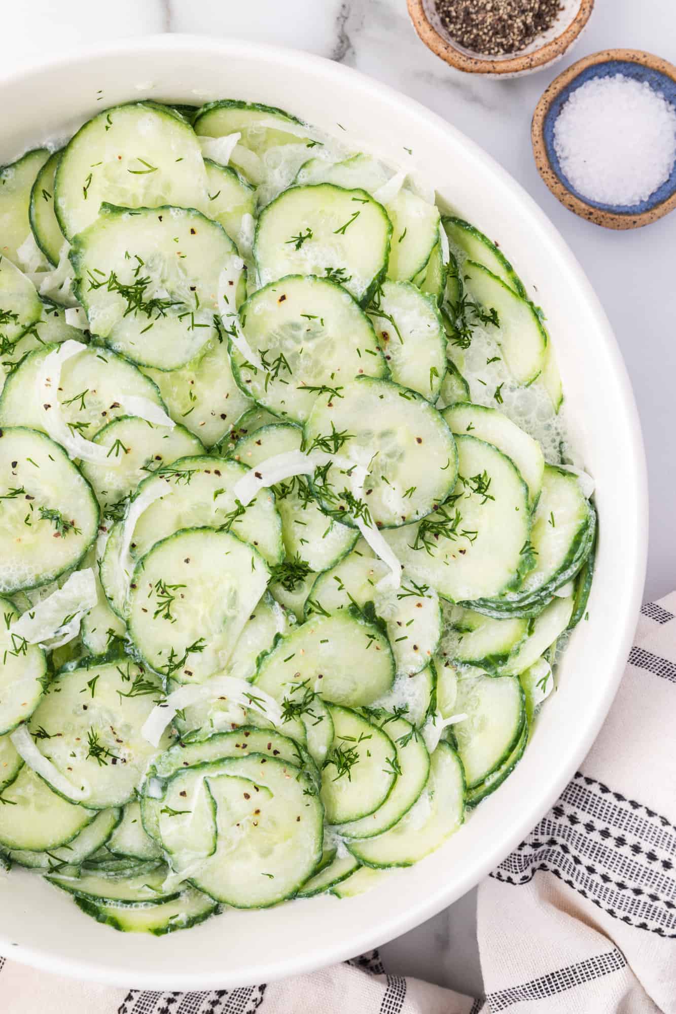 Overhead view of German cucumber salad in a white bowl, next to small ramekins of salt and pepper.