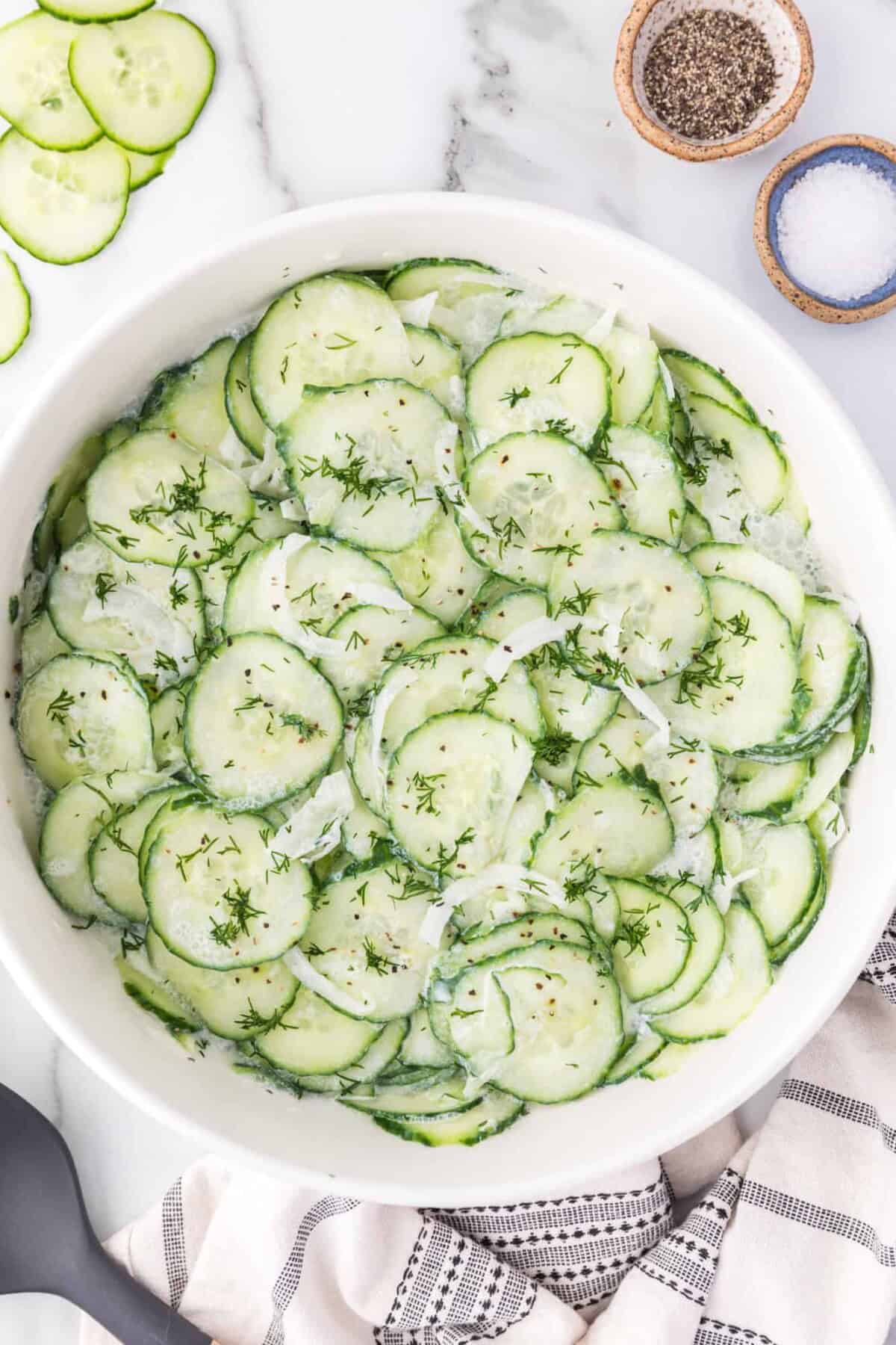 Overhead view of German cucumber salad in a white bowl, next to small ramekins of salt and pepper.