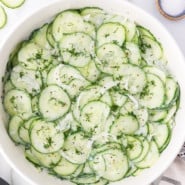 Overhead view of German cucumber salad in a white bowl, next to small ramekins of salt and pepper.