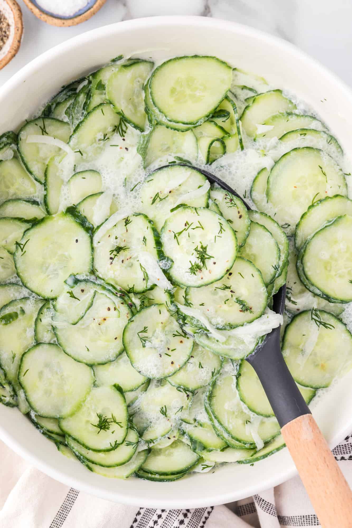 Overhead view of German cucumber salad in a white bowl with a serving spoon.