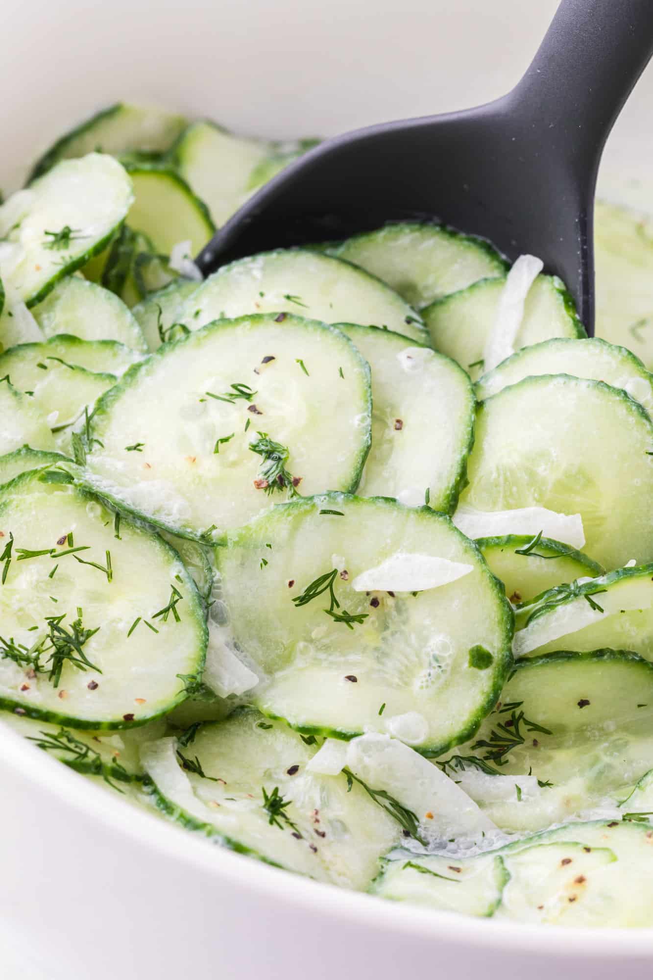 A black serving spoon scooping German cucumber salad from a large white bowl.