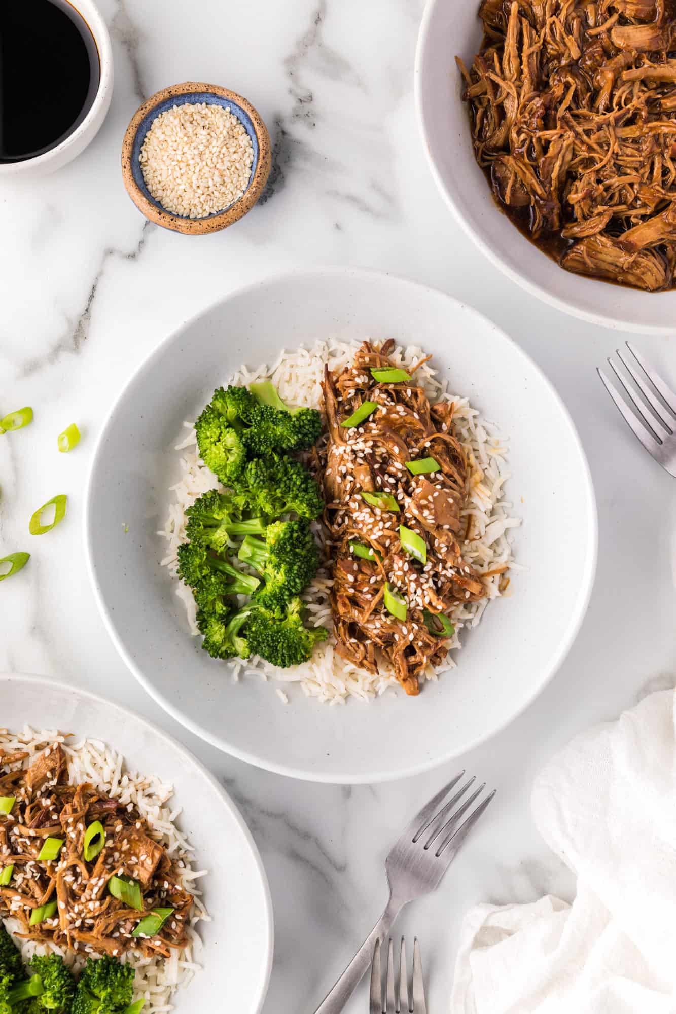 Overhead view of instant pot teriyaki chicken and steamed broccoli served over a bed of rice on plates, next to a bowl of shredded chicken teriyaki.