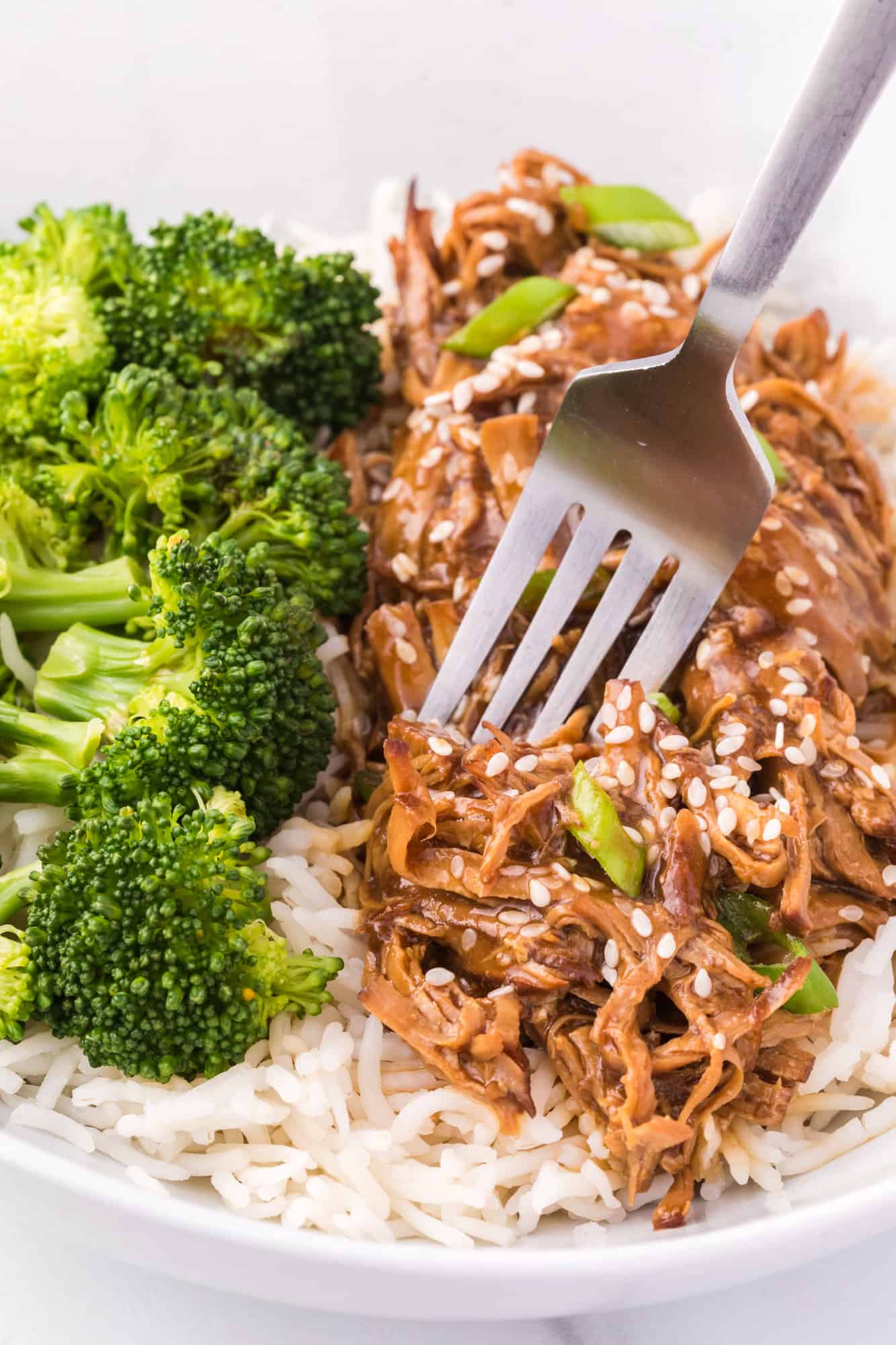 Close up of a fork picking from a pile of chicken teriyaki served next to broccoli in a bowl of white rice.