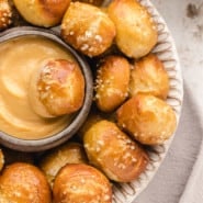 Overhead view of a pretzel bite dipped in a bowl of cheese sauce, surrounded by more pretzel bites on a white plate.