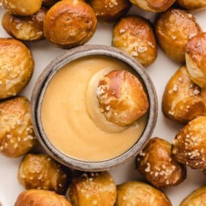 Overhead view of a pretzel bite dipped in a bowl of cheese sauce, surrounded by more pretzel bites on a white plate.