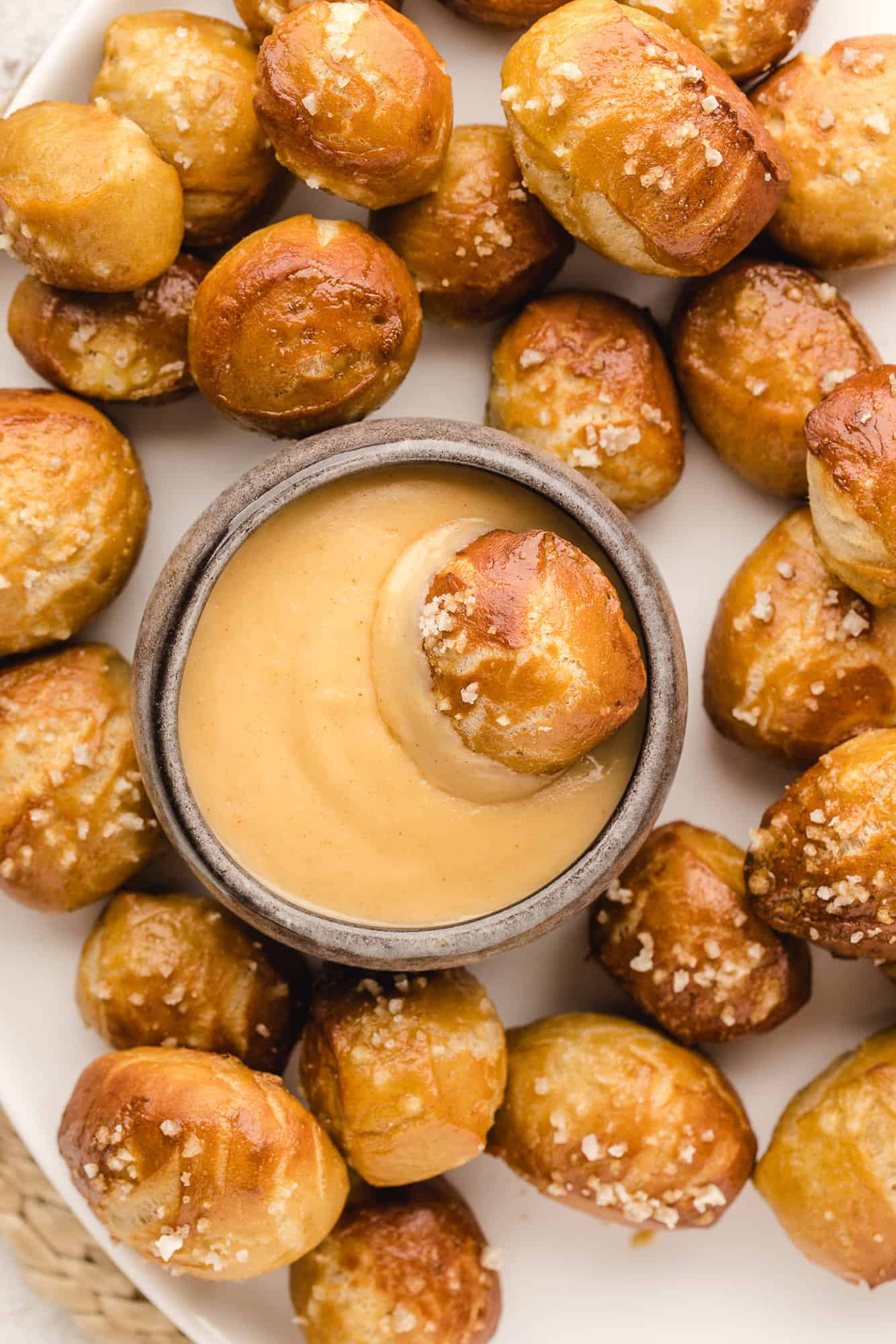 Overhead view of a pretzel bite dipped in a bowl of cheese sauce, surrounded by more pretzel bites on a white plate.