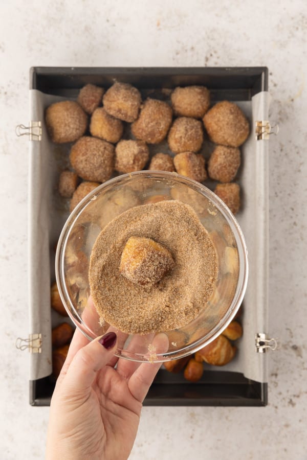 A hand holding a bowl of cinnamon sugar with a partially coated pretzel bite, over more pretzel bites on a baking sheet.
