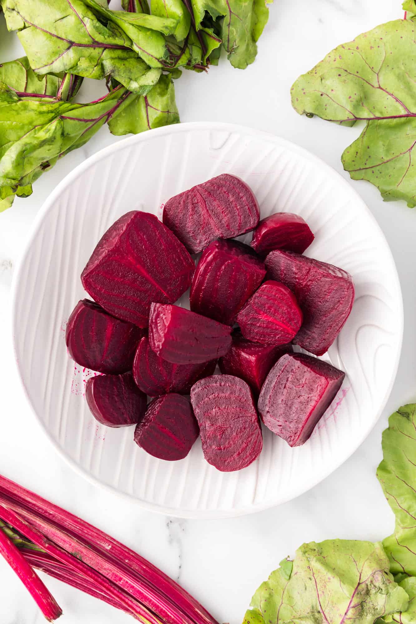 Chopped beets in a white bowl, surrounded by beet greens.