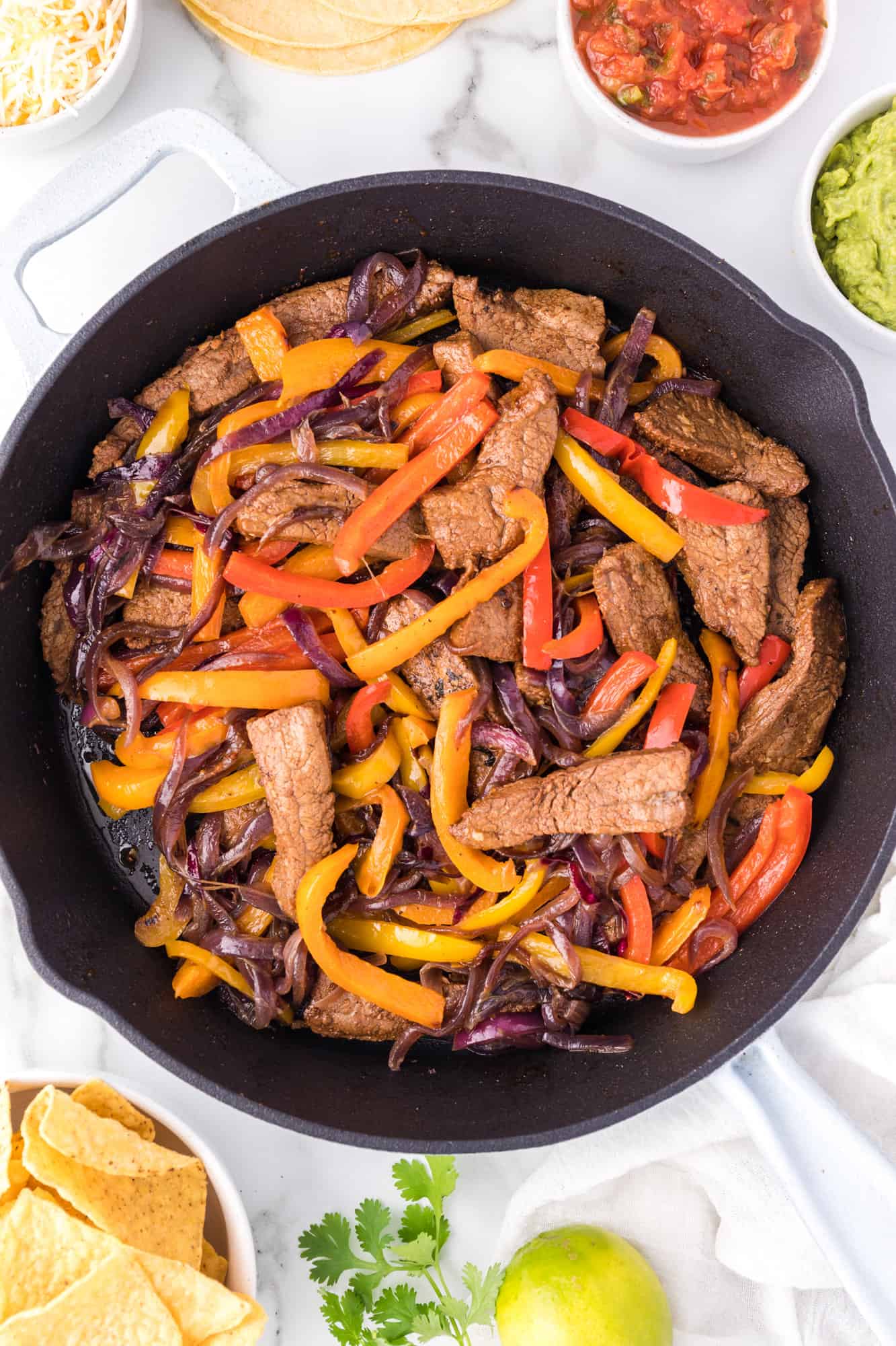 Overhead view of steak fajitas in a skillet, surrounded by bowls of toppings.