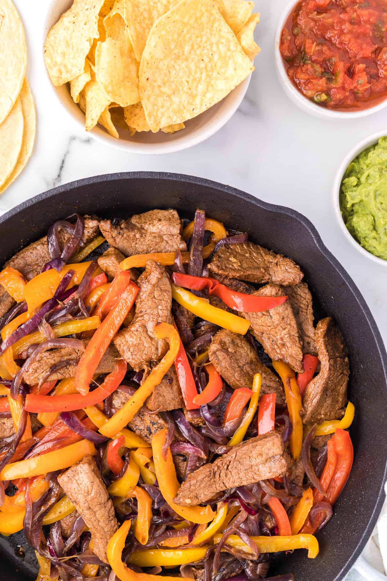Overhead view of steak fajitas in a skillet, next to bowls of tortilla chips, salsa, and guacamole.