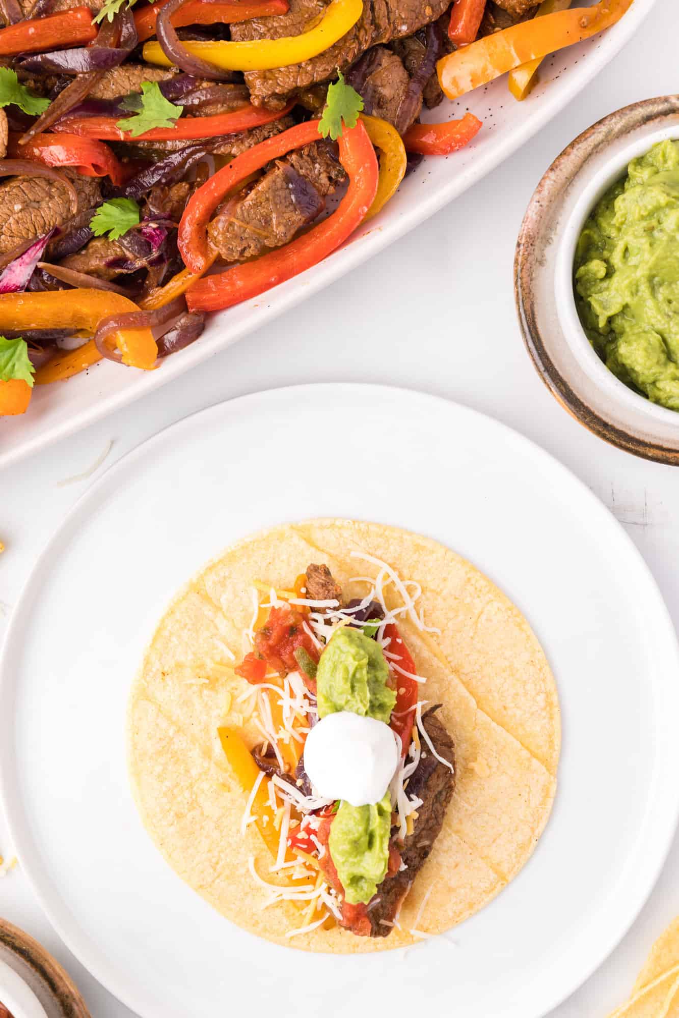 Overhead view of a tortilla topped with steak fajitas and garnishes on a plate, next to steak fajitas in a serving dish.