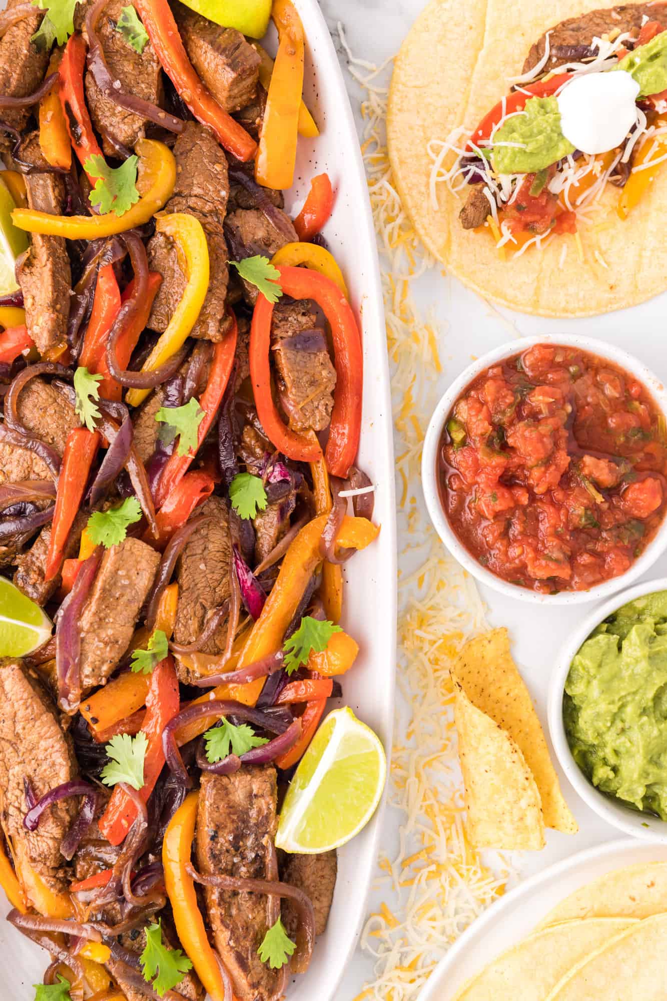 Overhead view of steak fajitas in a long serving dish, topped with lime wedges and cilantro, next to bowls of toppings and steak fajitas served over a tortilla on a plate.