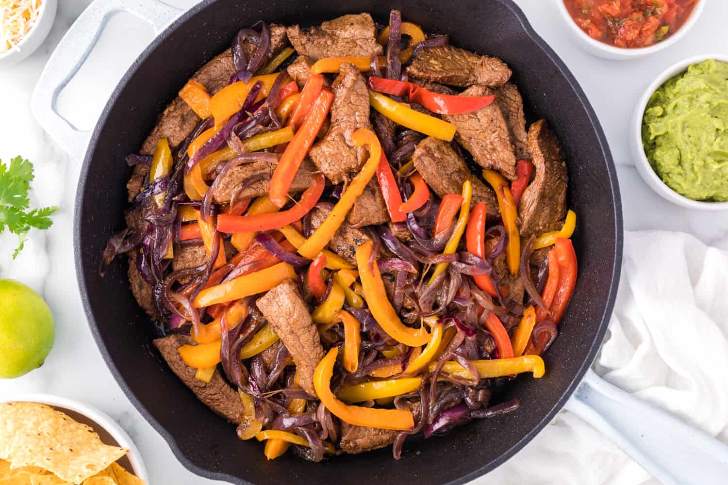 Overhead view of steak fajitas in a skillet, next to bowls of tortilla chips, salsa, and guacamole.