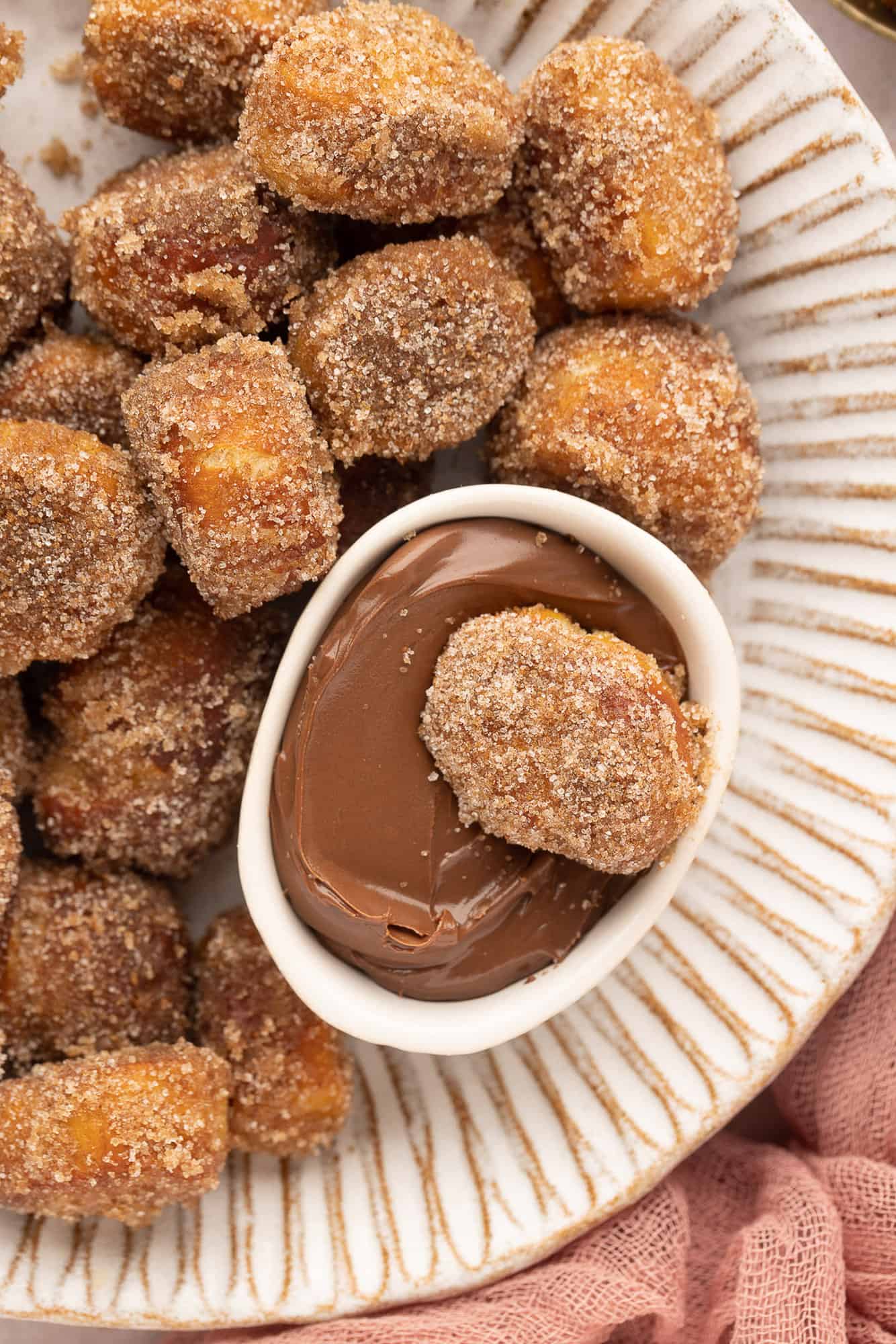 Close up overhead view of a cinnamon sugar pretzel bite resting in a small bowl of nutella, nestled in a larger bowl of pretzel bites.
