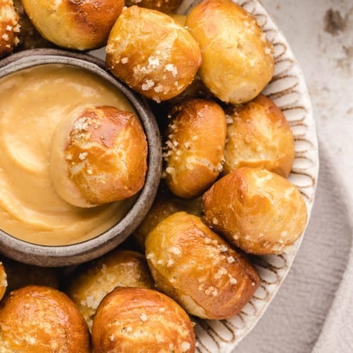 Overhead view of a pretzel bite dipped in a bowl of cheese sauce, surrounded by more pretzel bites on a white plate.