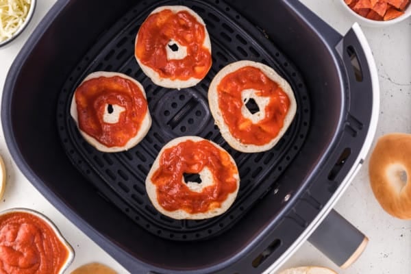 Mini bagel halves in air fryer, topped with pizza sauce.