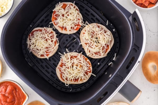 Mini bagel halves in air fryer, topped with pizza sauce and mozzarella.