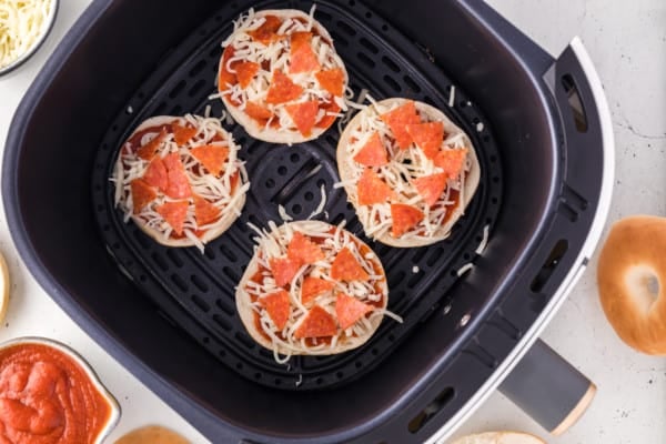 Mini bagel halves in air fryer, topped with pizza sauce, mozzarella, and pepperoni.