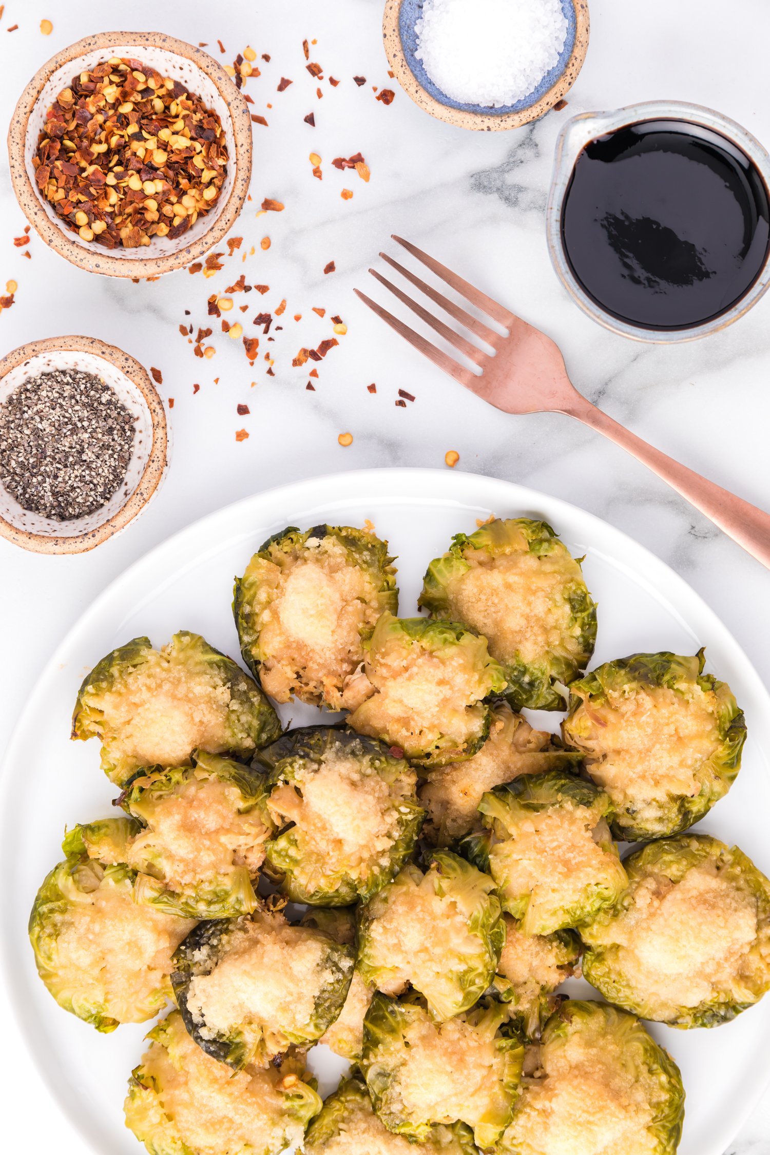 Cheese topped smashed Brussels sprouts, seasonings in small bowls nearby.