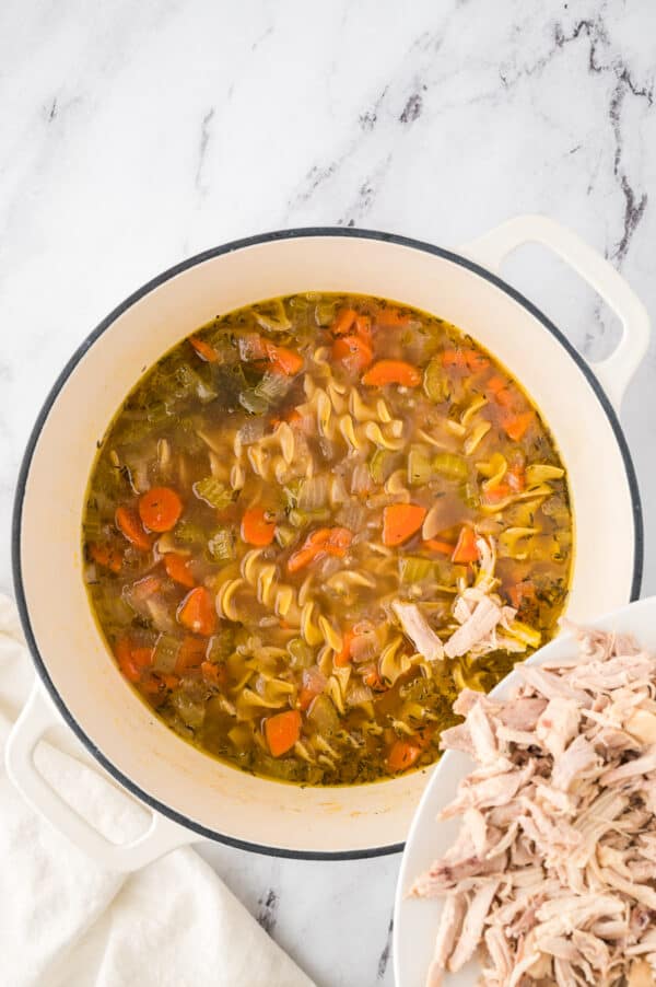 Shredded chicken being poured into a pot of chicken noodle soup.