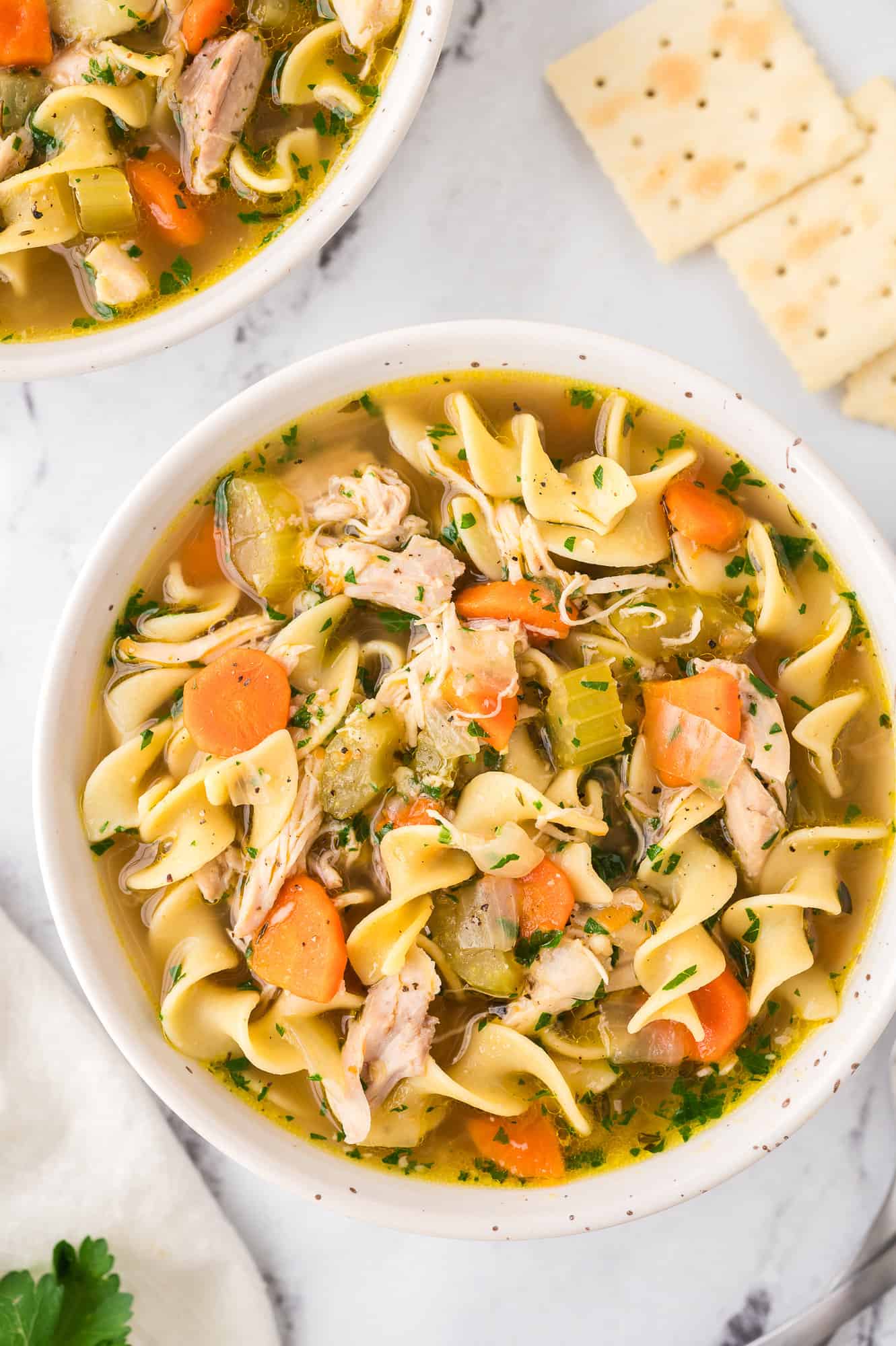 Overhead view of a bowl of chicken noodle soup next to a second bowl and saltine crackers.