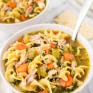 A bowl of chicken noodle soup with a second bowl of soup next to saltine crackers in the background.