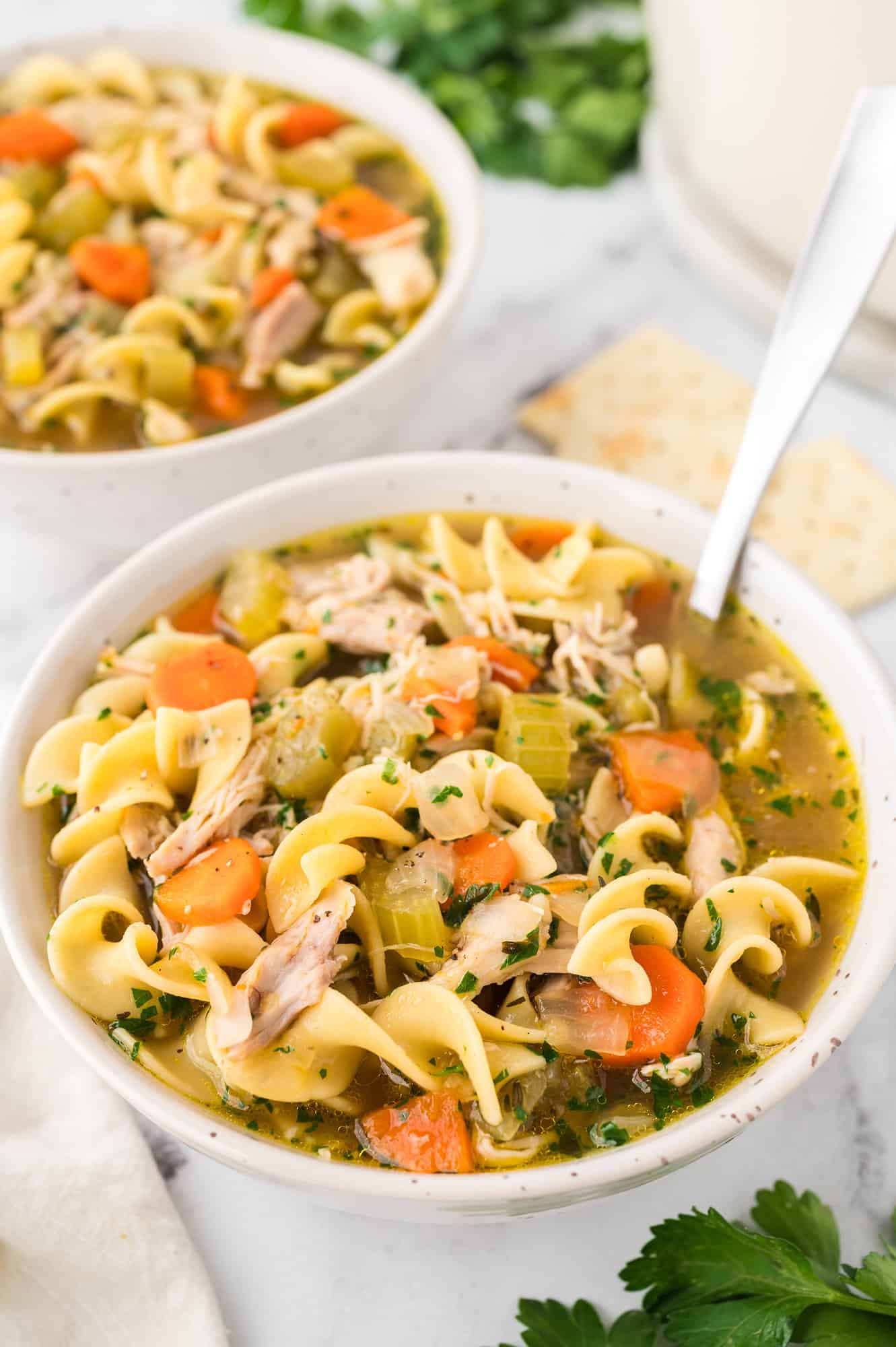 A bowl of chicken noodle soup with a second bowl of soup next to saltine crackers in the background.