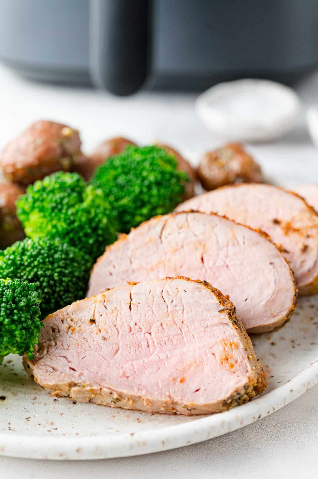 Plated pork tenderloin slices with broccoli, placed in front of an air fryer.